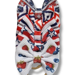 May include: Three fabric hair bows in patriotic red, white, and blue patterns. The top bow features a geometric design, the middle bow has stars and stripes, and the bottom bow showcases strawberries and blueberries. Each bow is attached to a tan elastic band.