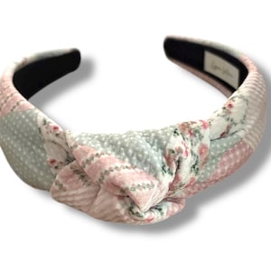 May include: A knotted headband with a pink and gray floral pattern. The headband is made of fabric and has a black elastic band.