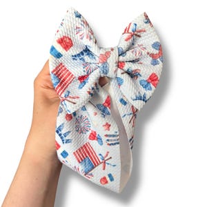 May include: White fabric bow with a patriotic print featuring red, white, and blue fireworks, flags, and stars. The bow is held in a hand and is designed for hair accessories.