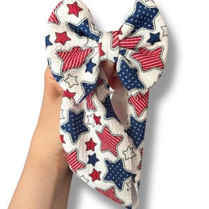 May include: A large white bow with a patriotic star pattern in red and blue. The bow is held up against a white background. The bow is made of a textured fabric.