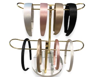 May include: A gold metal stand with two tiers for displaying headbands. The stand has a white base and holds eight headbands in various colors, including black, white, pink, and gold.
