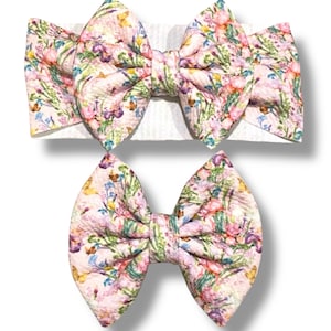 May include: Two floral print hair bows. The larger bow is on a white headband. The smaller bow is separate. Both bows have a pink background with green leaves and colorful flowers.