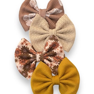 May include: Set of three hair bows in fall colors. The bows are made of fabric and have different patterns. One bow is brown with a white floral pattern, one is beige with a waffle texture, and one is mustard yellow with a waffle texture.