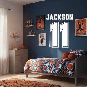 Könnte beinhalten: Ein Schlafzimmer mit Basketball-Thema. Die marineblaue Wand zeigt den Namen "JACKSON" über der Nummer "11" in Weiß. Ein Bett mit einer Bettdecke und Kissen im Basketball-Design befindet sich vor der Wand. Ein Basketballkorb und eine Kommode sind ebenfalls vorhanden.