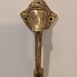 May include: A gold-toned metal door handle shaped like the head of the Hindu deity Ganesha. The handle has a detailed design and a long, curved spout.