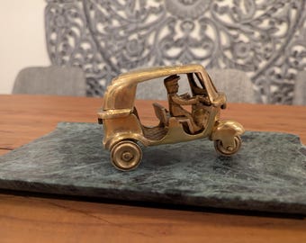 May include: A brass figurine of a three-wheeled auto rickshaw, also known as a tuk-tuk, with a driver figure inside. The rickshaw is on a green marble base.