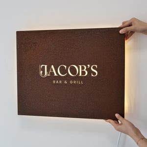 May include: A rectangular brown sign with the illuminated text "JACOB'S BAR & GRILL". The sign has a textured surface and is being held by two hands. The sign has a warm, inviting glow.
