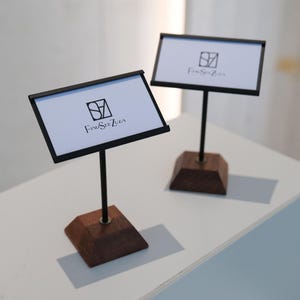 May include: Two display stands with black frames and dark wood bases. Each stand holds a white card with the logo "FindSeeZula" in black. The stands are on a white surface.