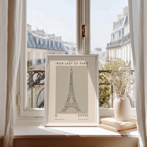 May include: A framed print of the Eiffel Tower, labeled "Iron Lady of Paris." The artwork features a minimalist line drawing of the tower in black on a beige background. The print is displayed on a windowsill with a view of Parisian buildings.