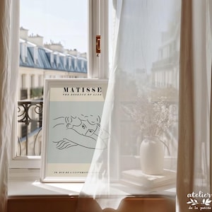 May include: A framed Matisse art print with a minimalist line drawing of two figures, displayed on a windowsill. A white vase with flowers and a stack of books are next to the print. Sheer white curtains frame the window, with a view of buildings.