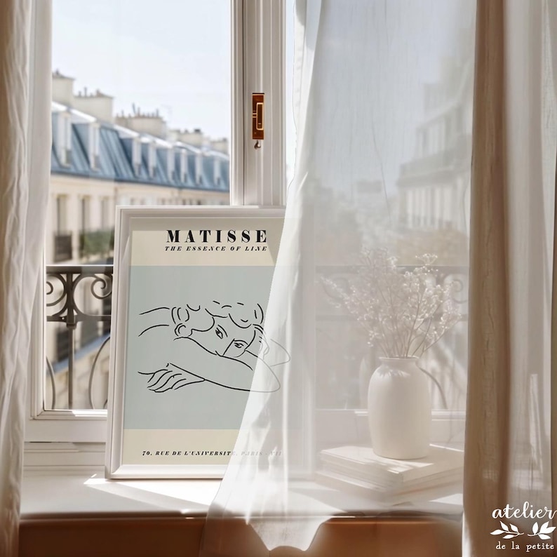 May include: A framed Matisse art print with a minimalist line drawing of two figures, displayed on a windowsill. A white vase with flowers and a stack of books are next to the print. Sheer white curtains frame the window, with a view of buildings.