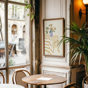 Könnte beinhalten: Ein gerahmter Kunstdruck mit einem Stillleben von Orangen in einer blau-weiß gestreiften Vase auf einer gelb-weiß gestreiften Tischdecke. Das Kunstwerk hängt in einem Café, mit einem Holztisch und Stühlen im Vordergrund.