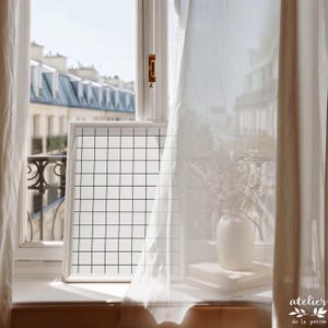 May include: A white framed grid art print sits on a windowsill, next to a white vase with dried flowers and a stack of books. Sheer white curtains frame the view of a city with buildings.