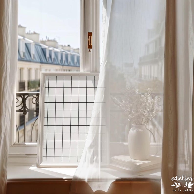 May include: A white framed grid art print sits on a windowsill, next to a white vase with dried flowers and a stack of books. Sheer white curtains frame the view of a city with buildings.