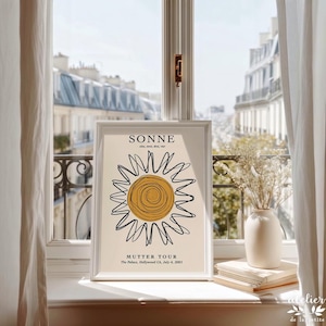 May include: A framed art print featuring a minimalist sun design with the word "SONNE" at the top. The print includes the text "MUTTER TOUR" and other details. The artwork is displayed on a windowsill with a view of a city.