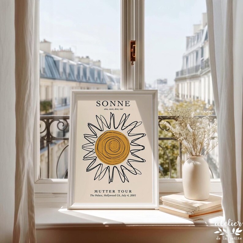 May include: A framed art print featuring a minimalist sun design with the word "SONNE" at the top. The print includes the text "MUTTER TOUR" and other details. The artwork is displayed on a windowsill with a view of a city.