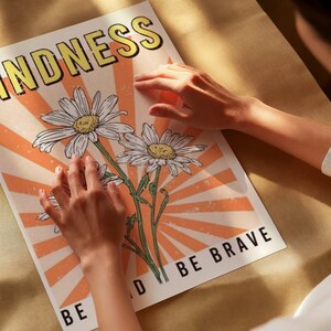May include: A motivational poster with a peach and white color scheme. The poster features a stylized illustration of three white daisies with yellow centers. The text on the poster reads "Kindness" and "Be Kind Be Brave".