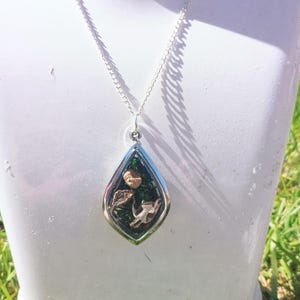 May include: Silver pendant necklace featuring a teardrop-shaped charm. The charm showcases a miniature scene with green, brown, and beige details, including a small figurine. The necklace is displayed on a white mannequin.