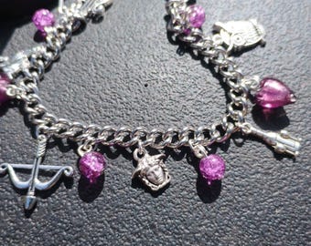 May include: A silver-toned charm bracelet featuring a curb chain with various dangling charms, including a bow and arrow, a Medusa head, an owl, a lightning bolt, and several translucent purple crackle glass beads interspersed along the metal links.