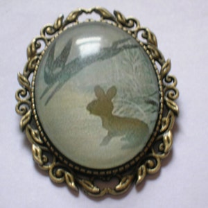 May include: A bronze-coloured metal brooch with a glass cameo featuring a stylised image of a rabbit and a bird in flight. The cameo is set in a decorative frame with a floral design.