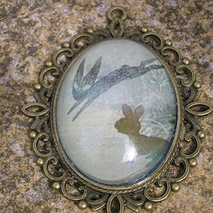 May include: An oval-shaped pendant made of bronze with an ornate design. The pendant features a glass cameo with a vintage illustration of a bird and a rabbit.