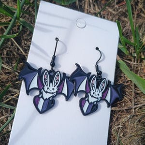 May include: A pair of black bat-shaped earrings with purple and white accents. The earrings feature a smiling bat design with heart-shaped bodies and black ear hooks. The earrings are displayed on a white card against a backdrop of green grass.