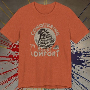 Może przedstawiać: Pomarańczowy T-shirt z vintage'ową grafiką osoby wspinającej się na stos poduszek z tekstem "Conquering Mount Comfort".