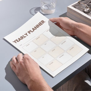 Könnte beinhalten: Ein weißer Jahresplaner mit dem braunen Titel "YEARLY PLANNER" und einem Raster mit 12 Monaten, von Januar bis Dezember.