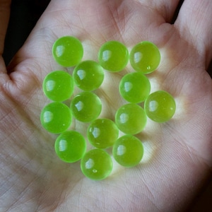 Puede incluir: Una colección de 12 canicas de vidrio verde, algunas con un ligero tono amarillo, están esparcidas en una mano.