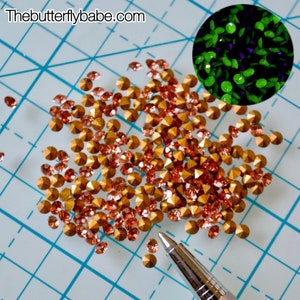 Può includere: Un mucchio di piccoli strass dorati con un luccichio color pesca. Gli strass sono sparsi su una superficie blu a griglia. Una penna d'argento giace sulla superficie accanto agli strass. Il sito web "Thebutterflybabe.com" è visibile nell'angolo in alto a sinistra dell'immagine.