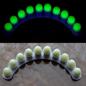 Può includere: Un set di dieci sfere verdi luminose disposte ad arco su sfondo nero. Sotto, le stesse sfere sono giallo pallido, ciascuna su un supporto grigio all'interno di un supporto curvo grigio, su una superficie di legno.