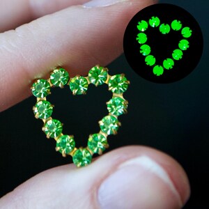 Puede incluir: Un broche en forma de corazón hecho de metal dorado con diamantes de imitación verdes. Los diamantes de imitación brillan en la oscuridad.