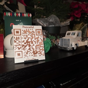 May include: A white card with a copper-colored QR code and the word "Peaceful" is displayed on a black stand. A small white truck with a Christmas tree in the back is to the right. A Christmas tree and poinsettias are in the background.