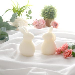 May include: Two white bunny shaped candles on a white fabric surface with pink flowers and green plants in the background.
