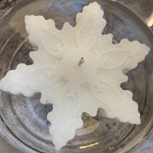May include: A white snowflake-shaped candle with glitter embedded in the wax. The candle is floating in a clear glass container.