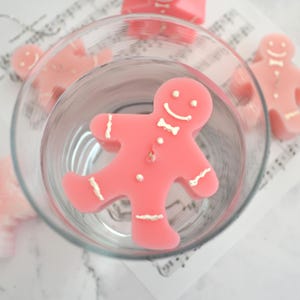 May include: A pink gingerbread man candle floating in a glass bowl of water. The candle has a white bow tie and a smiling face.