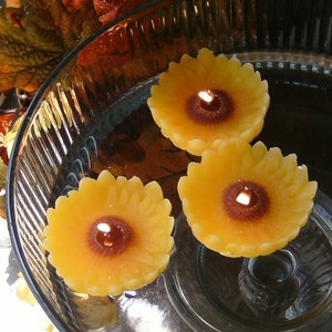 May include: Three yellow sunflower-shaped candles floating in a glass bowl filled with water. The candles have a brown center and are lit.