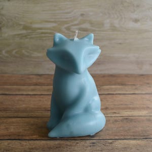 May include: A light blue fox-shaped candle with a white wick. The candle is sitting on a wooden surface.