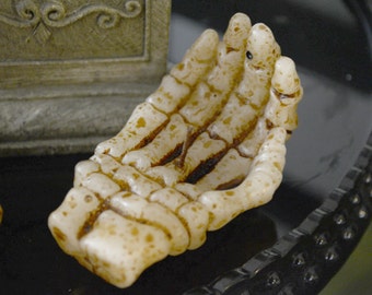 May include: A white skeletal hand candle holder with brown detailing. The hand is cupped, as if offering something.