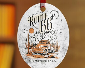 Op de afbeelding: Een ronde, witte, metalen ornament met een vintage Route 66-ontwerp. De ornament toont een rode pick-up truck die door een woestijnlandschap met cactussen en bergen rijdt. De tekst "Route 66" is in oranje en zwart, en de tekst "The Mother Road" is in zwart.
