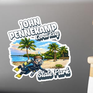 Op de afbeelding: Een sticker met een cartoon illustratie van een duiker bij een strand met palmbomen. De sticker heeft de tekst "John Pennekamp Coral Reef State Park" in een witte, gestileerde letter.