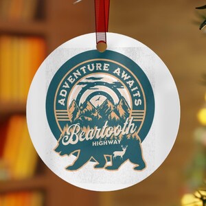 May include: A round white ornament with a green and gold design. The ornament features a bear walking in front of a mountain range with the text "Adventure Awaits Beartooth Highway".