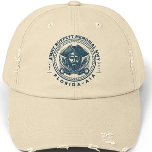 May include: A beige distressed baseball cap with a blue and white logo featuring a pirate with crossed swords and the text "Jimmy Buffett Memorial Hwy. Est. 2014 Florida - Aia"
