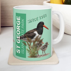 Peut inclure: Mug en céramique blanche avec une illustration d'oiseau, avec un bec rouge et des plumes noires et blanches, sur un fond vert clair. Le mug porte les inscriptions "ST GEORGE ISLAND STATE PARK" et "SINCE 1963".