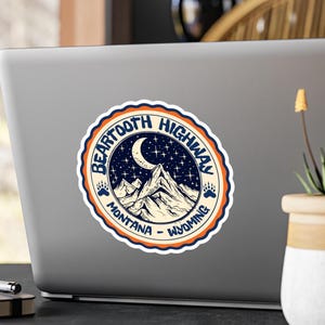 May include: A round sticker with a vintage design featuring the text "Beartooth Highway" and the states of Montana and Wyoming. The sticker has a blue and orange border, with a mountain range, moon, and stars in the center.