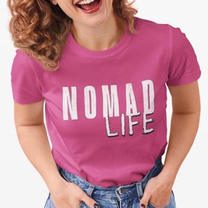 May include: A magenta t-shirt with the words "NOMAD LIFE" printed in white, with a black shadow effect. The shirt is being worn with blue jeans.