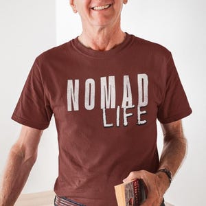 May include: A maroon t-shirt with the words "NOMAD LIFE" printed in a distressed white font. The shirt has short sleeves and a crew neck. The person is holding a book.