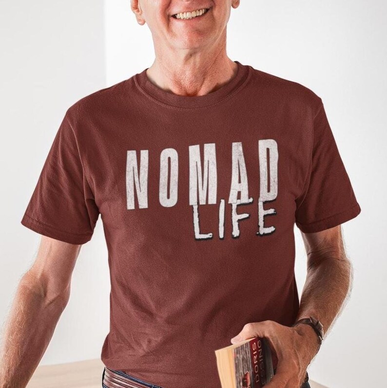 May include: A maroon t-shirt with the words "NOMAD LIFE" printed in a distressed white font. The shirt has short sleeves and a crew neck. The person is holding a book.