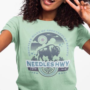 May include: A light green t-shirt featuring a circular graphic with a bison, the text "Needles Hwy," and the phrase "Fourteen Miles of Spectacular Beauty." The design includes stars, a sunburst, and the establishment year 1919.