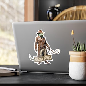 May include: A sticker featuring an iguana wearing a brown fedora and a brown leather jacket. The iguana is holding a whip and has the text "Iguana Jones" on a banner.